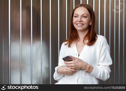 Thoughtful pleased woman with dreamy look, holds mobile phone, sends text message, wears white formal jumper, connected to wireless internet, uses new application. Lady with up to date digital device