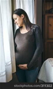 Thoughtful pregnant woman caressing her belly and looking out the window. Pregnant woman caressing belly and looking window