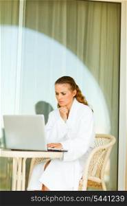 Thoughtful pretty woman in bathrobe sitting at table on terrace and using laptop