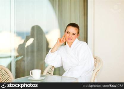 Thoughtful pretty woman in bathrobe sitting at table on terrace with cup of coffee&#xA;