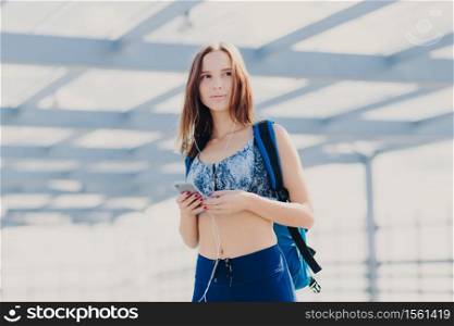 Thoughtful pretty young woman with healthy clean skin, dressed in casual top and leggings, has bag, holds modern electronic gadget, entertains herself with music, looks pensively into distance