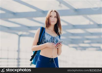 Thoughtful pretty young woman with healthy clean skin, dressed in casual top and leggings, has bag, holds modern electronic gadget, entertains herself with music, looks pensively into distance
