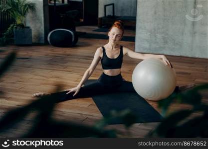 Thoughtful red haired woman in black sportswear sitting in split position on yoga mat in fitness studio and looking away from camera, enjoying pilates class. Healthy lifestyle and sport concept. Sportive woman relaxing during pilates training with exercise ball