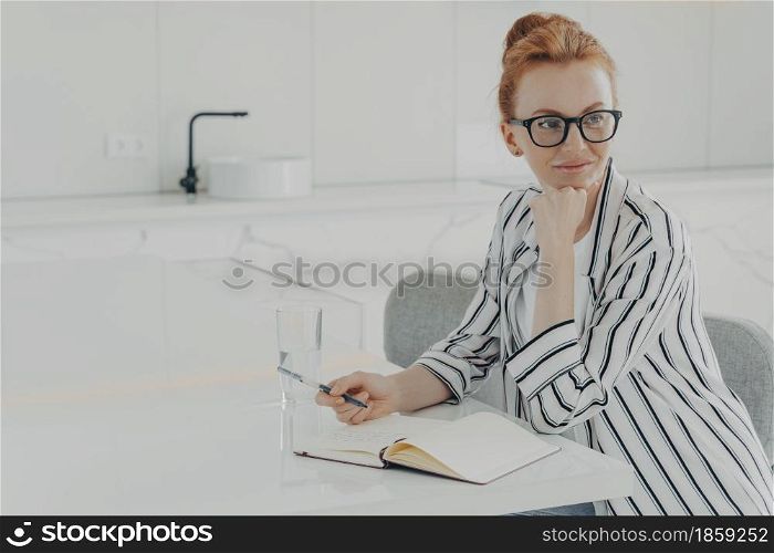 Thoughtful redhead woman distracted from work makes notes in notepad holds pen wears spectacles striped shirt poses at white desktop writes down information in notebook works distantly from home. Thoughtful redhead woman distracted from work makes notes in notepad holds pen