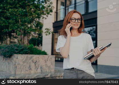 Thoughtful redhead woman looks into distance makes phone call holds smartphone near ear tablet and notebook wears white t shirt transparent glasses poses outdoor in urban place. Technology concept. Thoughtful redhead woman looks into distance makes phone call holds smartphone near ear