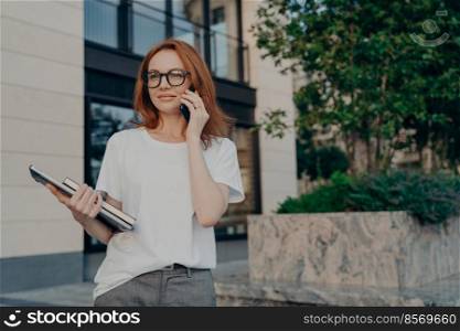 Thoughtful redhead woman looks into distance makes phone call holds smartphone near ear tablet and notebook wears white t shirt transparent glasses poses outdoor in urban place. Technology concept. Thoughtful redhead woman looks into distance makes phone call holds smartphone near ear