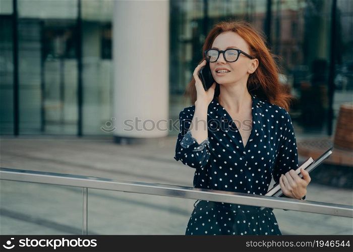 Thoughtful redhead woman wears spectacles polka dot dress has telephone conversation looks into distance stands near blurred office background carries necessary things for working. Lifestyle. Thoughtful redhead woman wears spectacles polka dot dress has telephone conversation