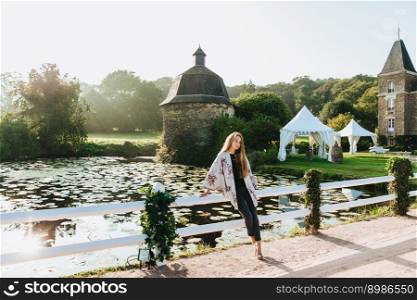 Thoughtful sad female in white cape stands near white short hence and small pond or lake, has walk on ancient historical territory, admires beautiful landscapes. People, lifestyle, excursion concept