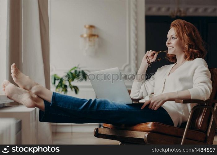 Thoughtful satisfied female freelancer works with computer at home, stretches legs and holds eyeglasses, searches information on social media sites, uses home internet connection for online chatting