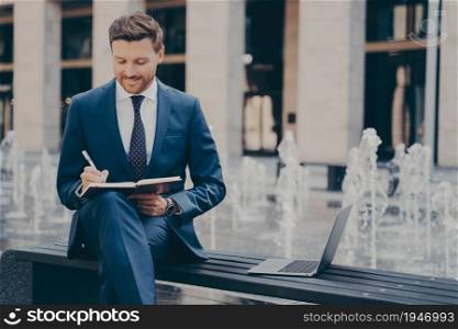 Thoughtful smiling young handsome professional entrepreneur in blue suit holding workbook and writing in it with pen, sitting on bench next to laptop, fountain in background behind him. Thoughtful smiling young businessman planning working day, making notes in notebook outdoors
