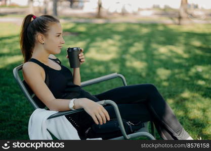 Thoughtful sporty young woman with pony tail has coffee break after training poses in comfortable chair with hot drink in park against green grass enjoys sunny day concentrated into distance