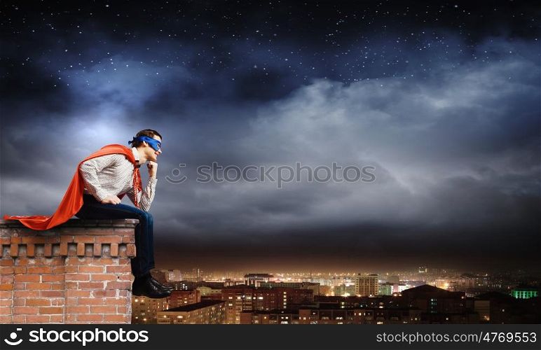 Thoughtful superman. Thoughtful superman in cape and mask sitting on top of building