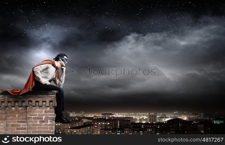 Thoughtful superman. Young man in superhero costume sitting on top of building