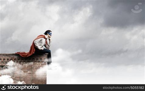 Thoughtful superman. Young man in superhero costume sitting on top of building