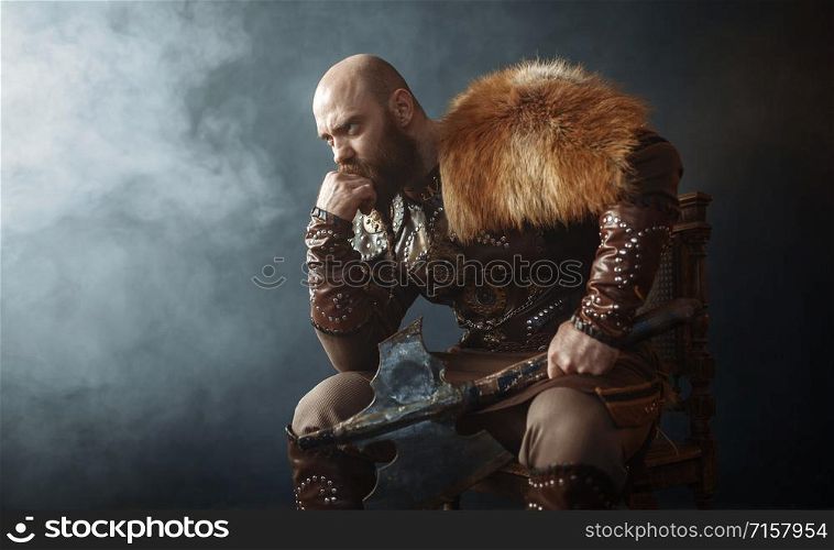 Thoughtful viking with axe dressed in traditional nordic clothes sitting on chair, barbarian image. Ancient warrior in smoke on dark background