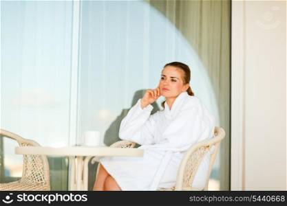 Thoughtful woman in bathrobe sitting at table on terrace