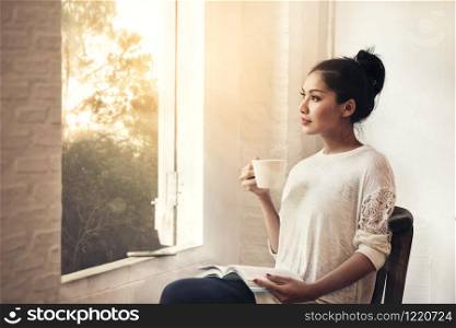 Thoughtful young beauty woman white cloth with book and cup of coffee sitting looking through the window, morning forrest landscape outside