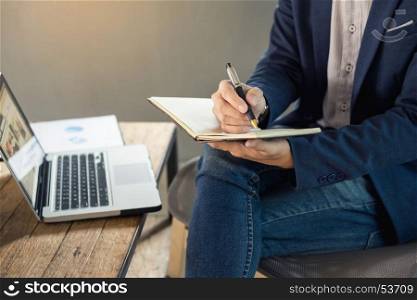 Thoughtful young business man in casual shirt holding note pad in a notebook on a wooden table with pen working in a cafe.