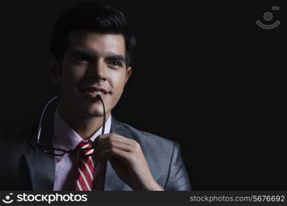 Thoughtful young businessman holding glasses against black background