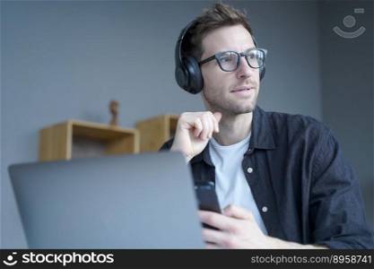 Thoughtful young businessman in headphones holds smartphone while sits at workplace at home office looking pensively aside, plunges into deep thoughts coming up with new ideas. Freelance concept. Thoughtful young businessman in headphones holds smartphone while sits at workplace at home