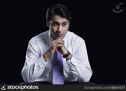 Thoughtful young businessman sitting with hands clasped over black background