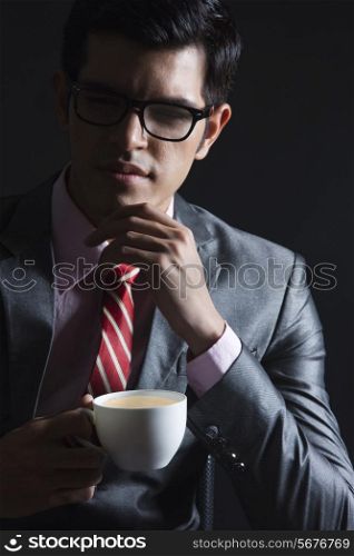 Thoughtful young businessman with coffee cup against black background