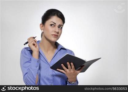 Thoughtful young businesswoman holding note pad and pen against gray background