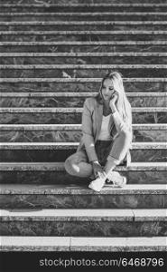 Thoughtful young caucasian woman in urban background. Blond girl wearing casual clothes in the street. Female with elegant jacket and blue jeans sitting on stairs.