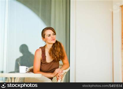 Thoughtful young female sitting on terrace with cup of tea and looking in corner&#xA;