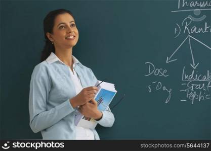 Thoughtful young female teacher holding books and glasses against green board