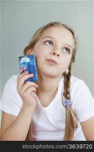 Thoughtful young girl holding credit card over gray background