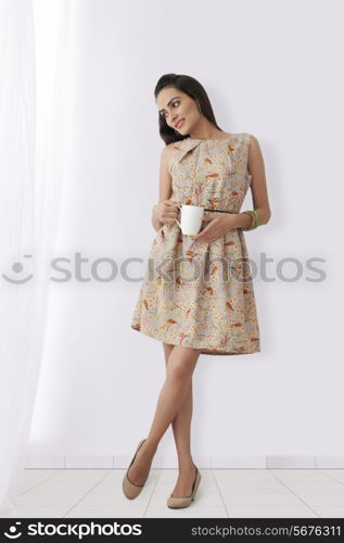 Thoughtful young Indian woman with coffee cup standing against white wall