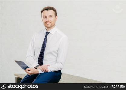 Thoughtful young male economist dressed in elegant luxury clothes, works on touch pad during break, poses in modern office corridor, thinks about cooperation, isolated over white background.