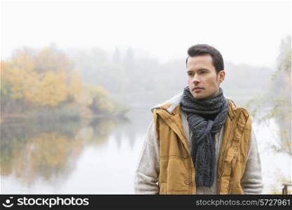 Thoughtful young man in warm clothing standing against lake