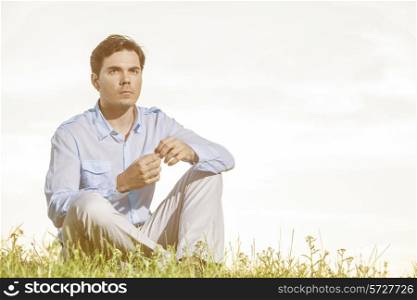 Thoughtful young man looking away while sitting on grass against clear sky