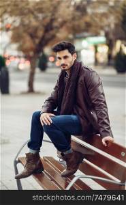 Thoughtful young man sitting on an urban bench. Attractive guy with modern hairstyle with lost look in the street.. Thoughtful young man sitting on an urban bench.