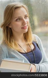 Thoughtful young student holding books by window during rainy day