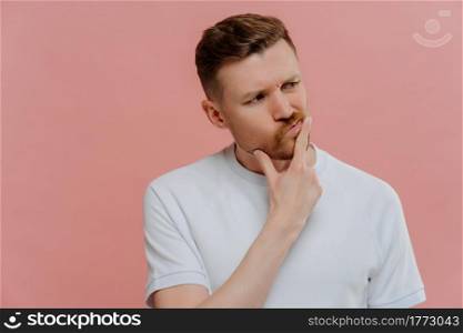 Thoughtful young unshaven man in white shirt having doubts and thinking about something with confused face expression while standing isolated over pink background. Hesitation concept. Young man having doubts, looking aside and thinking