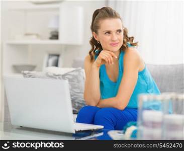 Thoughtful young woman sitting on couch and using laptop in living room