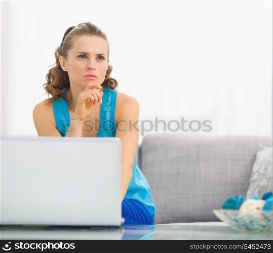 Thoughtful young woman sitting on divan and using laptop in living room