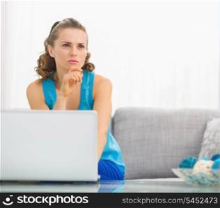 Thoughtful young woman sitting on divan and using laptop in living room