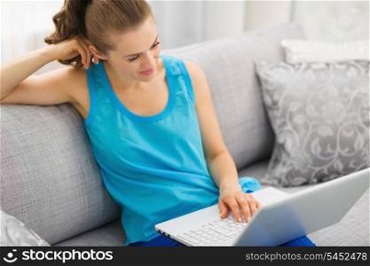 Thoughtful young woman sitting on sofa and using laptop in living room