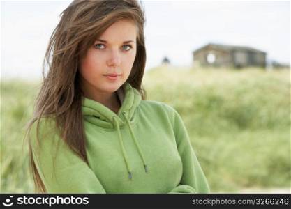 Thoughtful Young Woman Standing On Beach Wearing Hooded Top With Old Beach Hut In Distance