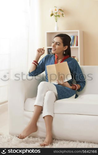Thoughtful young woman with book sitting on sofa