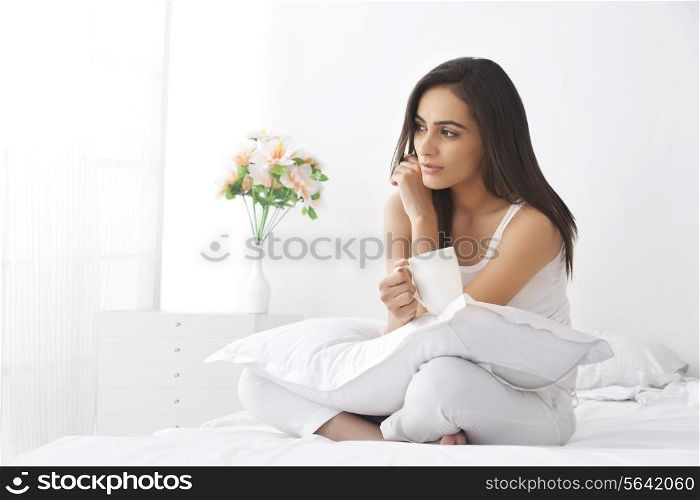 Thoughtful young woman with coffee cup sitting on bed