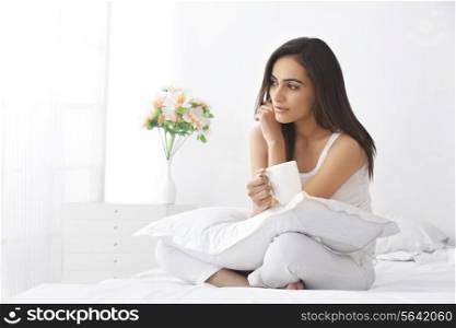 Thoughtful young woman with coffee cup sitting on bed