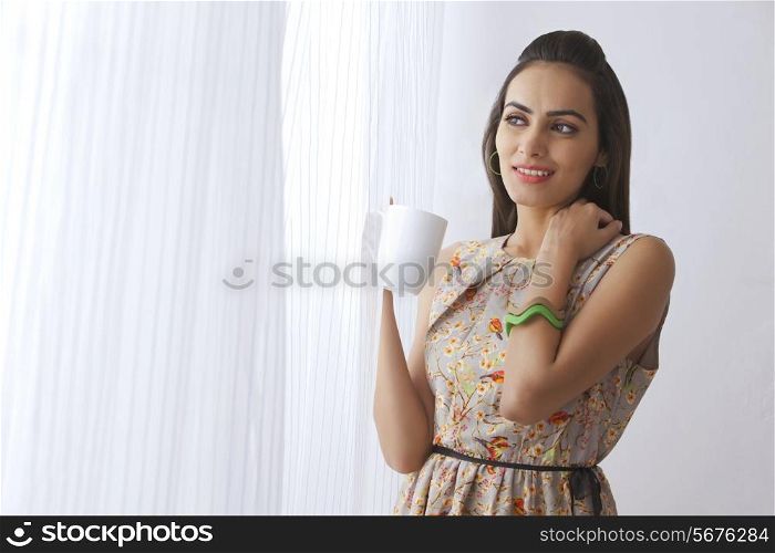 Thoughtful young woman with coffee cup standing by curtains