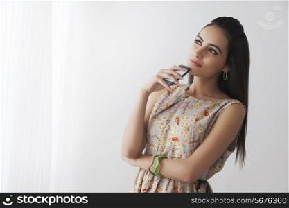 Thoughtful young woman with mobile phone standing by curtains