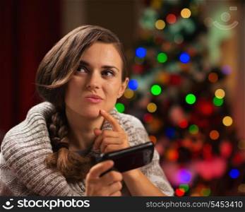 Thoughtful young woman writing sms in front of Christmas tree