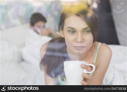 Thougtful woman having coffee at home with man lying in background